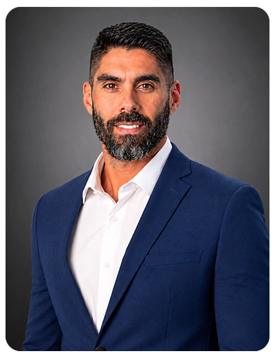 Chiropractor Rafael Sierra In Navy Suit Smiling In Front Of Grey Background