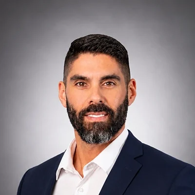 Chiropractor Rafael Sierra In Navy Suit Smiling In Front Of Grey Background