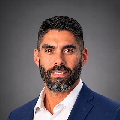 Chiropractor Rafael Sierra In Navy Suit Smiling In Front Of Grey Background